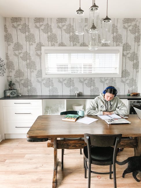 Focused young female student in warm hoodie and blue headsets reading notes in copybook while sitting on wooden table and doing homework in bright cozy kitchen